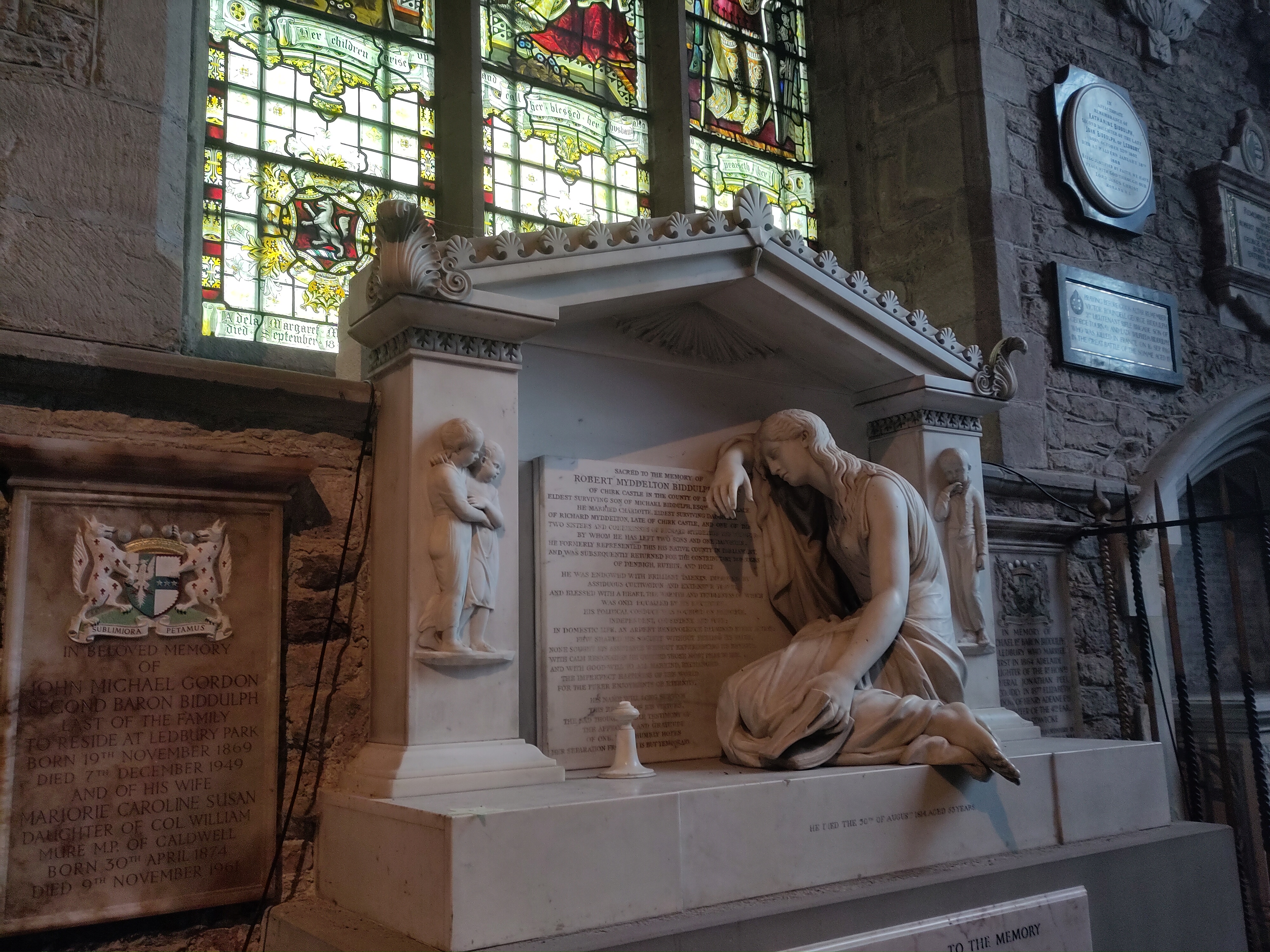 A photograph of the Biddulph memorial sculpture, directly underneath the window. It is made in a classical style using marble and depicts a mourning figure of a women, under a grecian canopy. She is leaning against a shield shape with is covered in an inscription, which is not legible from the photo.